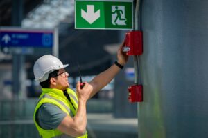 Monteur met helm en veiligheidshes test een rode handbrandmelder onder een groen nooduitgangsbord, terwijl hij via portofoon communiceert in een industriële hal.
