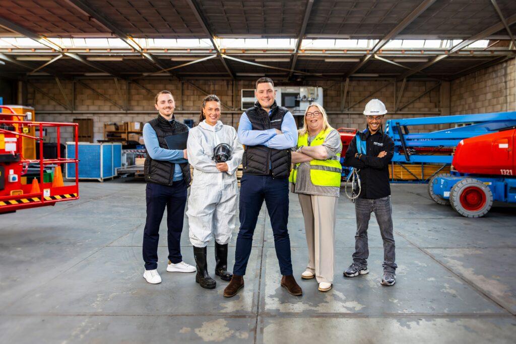 Team van Opleidingscentrum Westpoort poseert in praktijkloods voor veiligheid- en mobiliteitsopleidingen met hoogwerker op achtergrond.